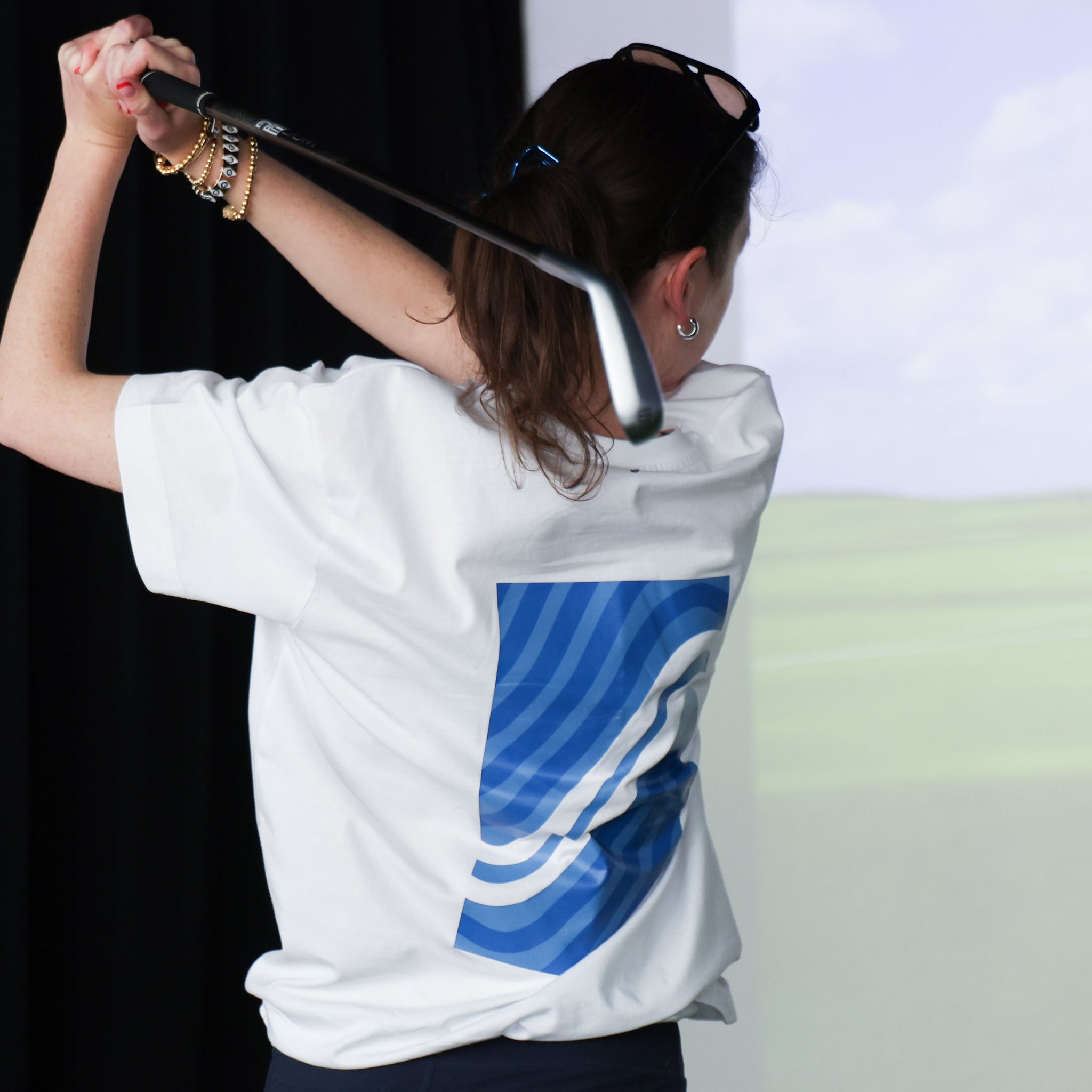 Woman wearing a white shirt with a blue logo, holding a golf club against a golf simulator.