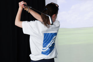 Woman wearing a white shirt with a blue logo, holding a golf club against a golf simulator.